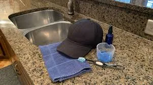 Black cap, towel, measuring cup, and soap on kitchen countertop near sink.