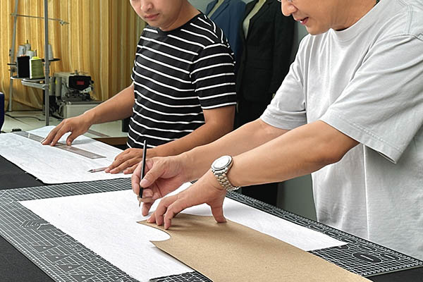 Two people drawing and cutting hat patterns on fabric table.