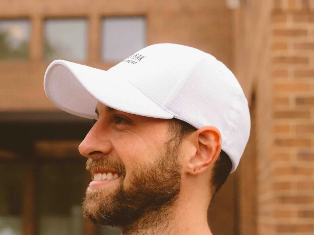 Smiling man with beard wearing white fitted cap outdoors.