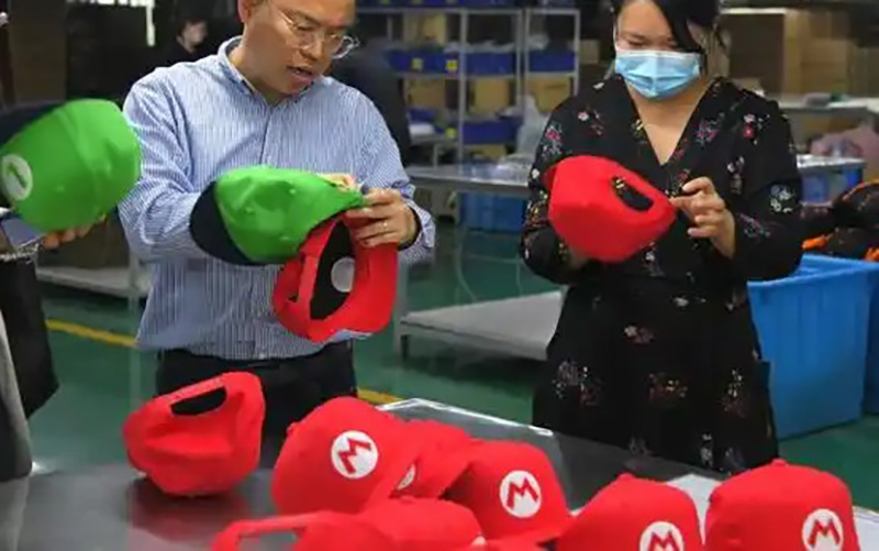 Two people inspecting and adjusting Mario-themed red and green caps in a production facility.
