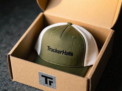 Olive green and white trucker hat with “TruckerHats” text in cardboard box.
