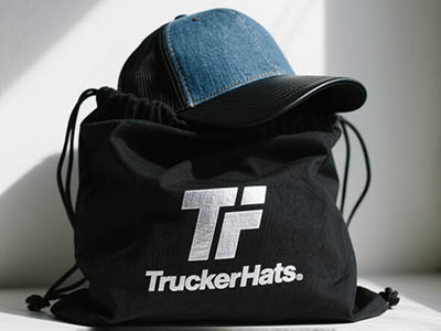 Denim and black trucker hat resting on black bag with “TruckerHats” logo.