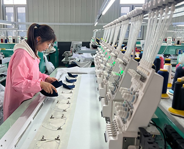 Worker inspecting caps near automated embroidery machines in factory.