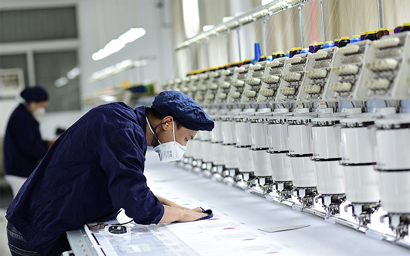 Man in mask inspecting fabric at embroidery machine in factory.