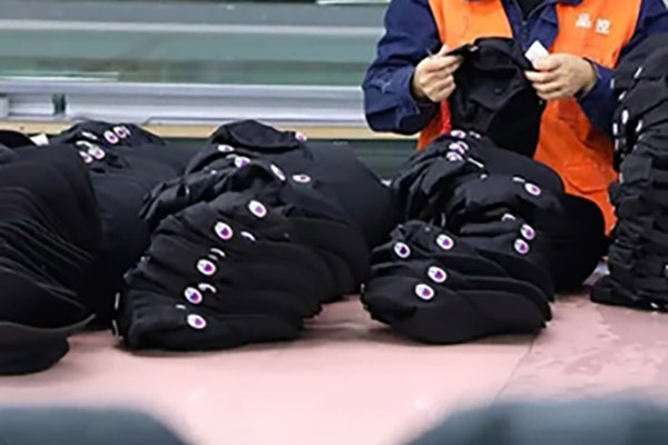 Worker inspecting black caps with embroidered logos in a factory setting.
