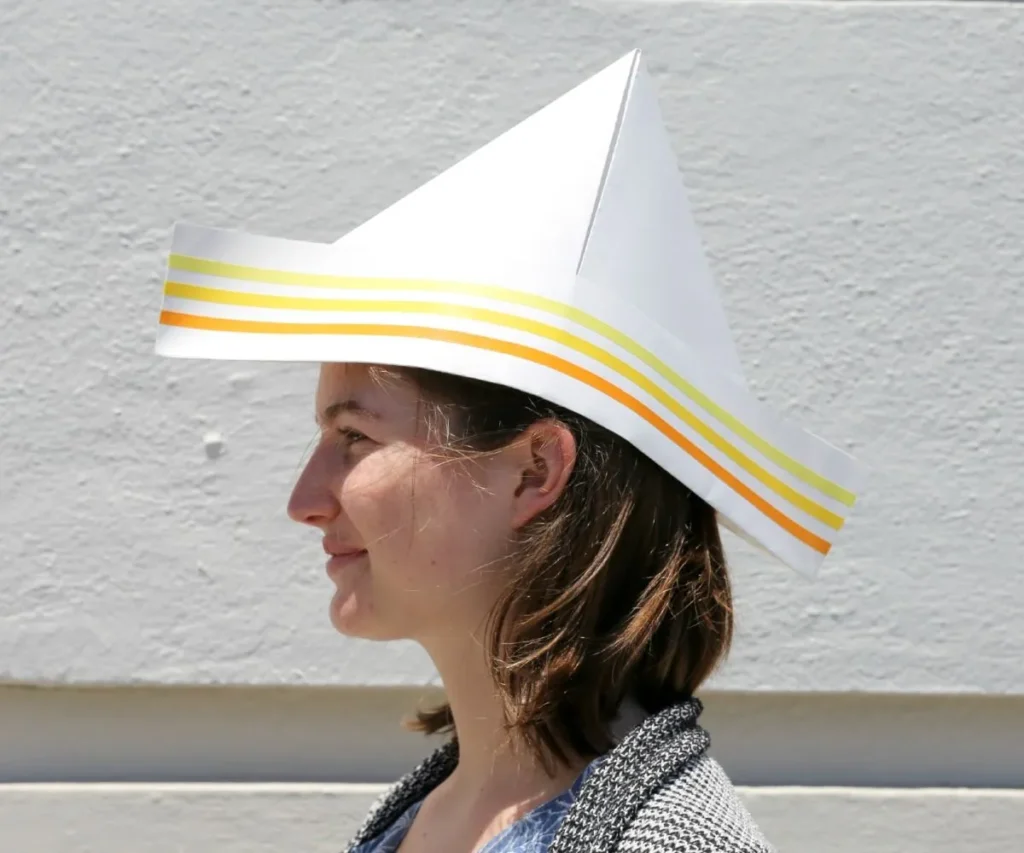 Side profile of woman wearing white paper hat with orange and yellow stripes