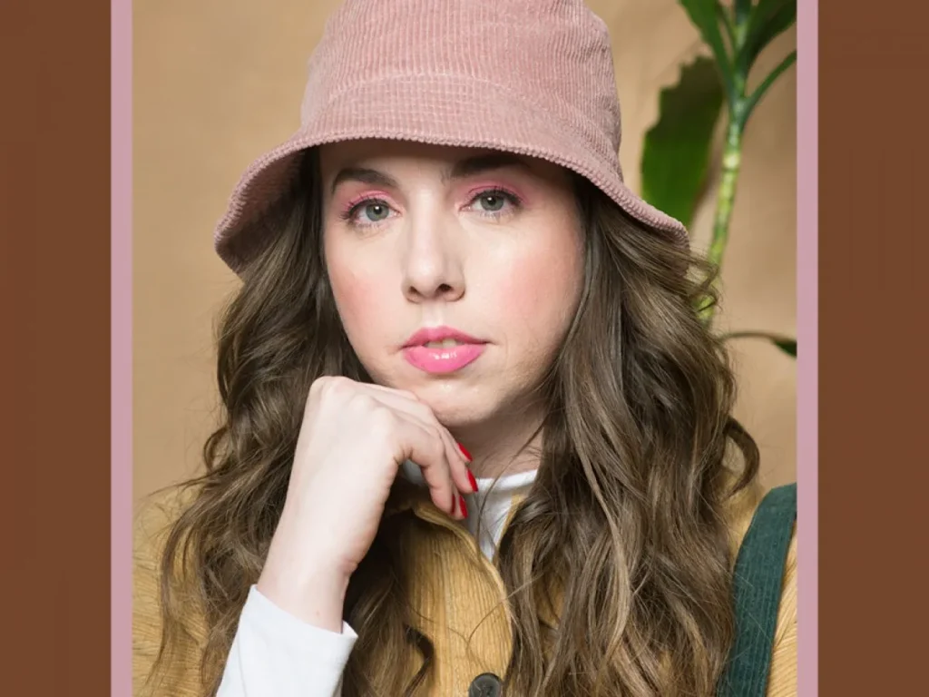 Woman with long curly hair wearing pink corduroy bucket hat and mustard jacket.