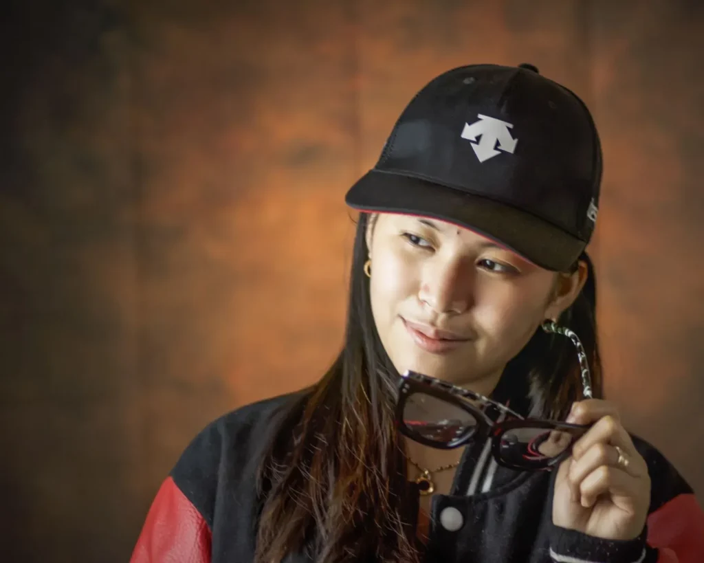 Woman in black jacket holding sunglasses, wearing black cap with white logo.