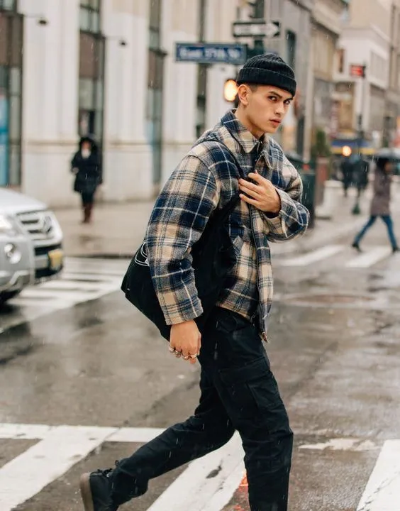 Young man wearing black beanie and plaid jacket walking on rainy city street